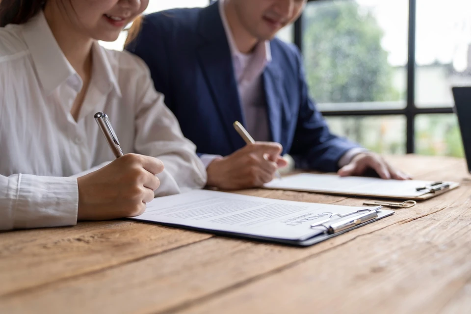 Deux professionnels en tenue d'affaires signent des documents contractuels dans un bureau moderne avec baie vitr&eacute;e