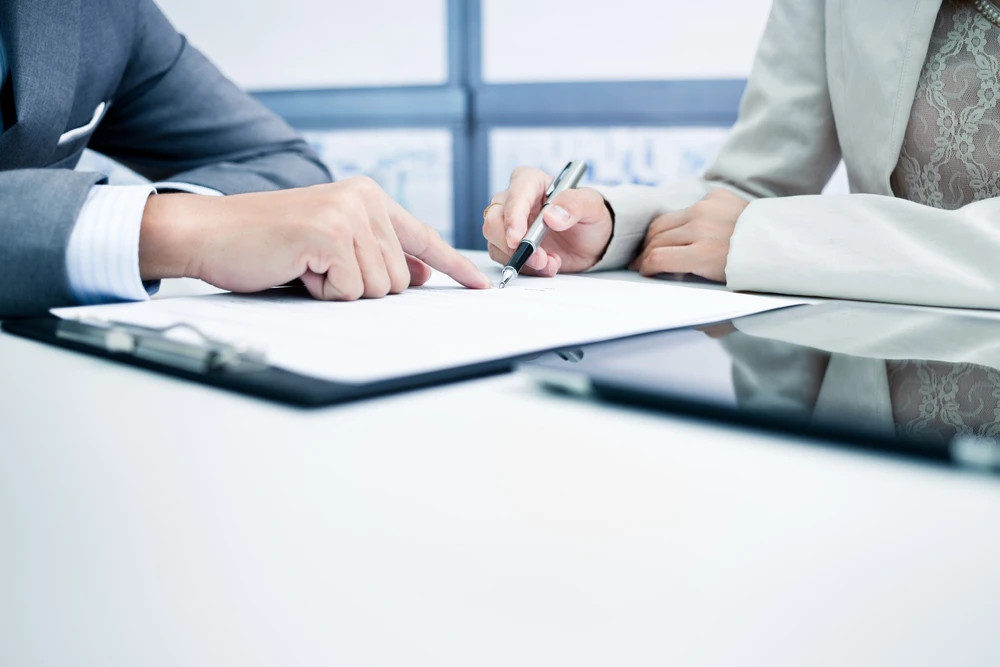 Scène de bureau professionnel avec deux personnes en costumes signant un document contractuel sur une table blanche