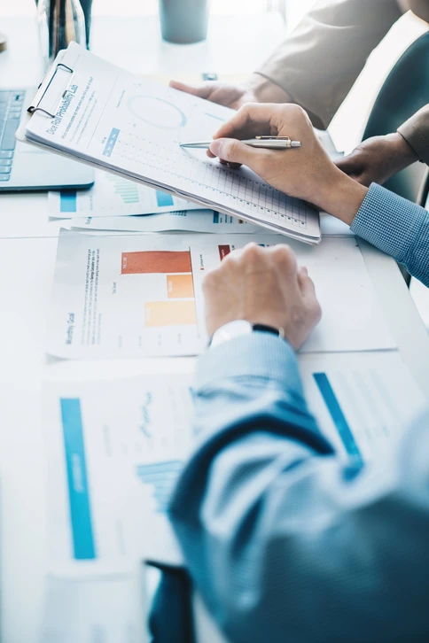 Scène de travail collaboratif en bureau avec deux professionnels analysant des documents financiers et graphiques colorés étalés sur table blanche