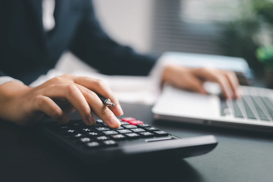 Personne en chemise bleue déposant une pièce dans un bocal en verre rempli de monnaie, calculatrice et documents financiers sur le bureau